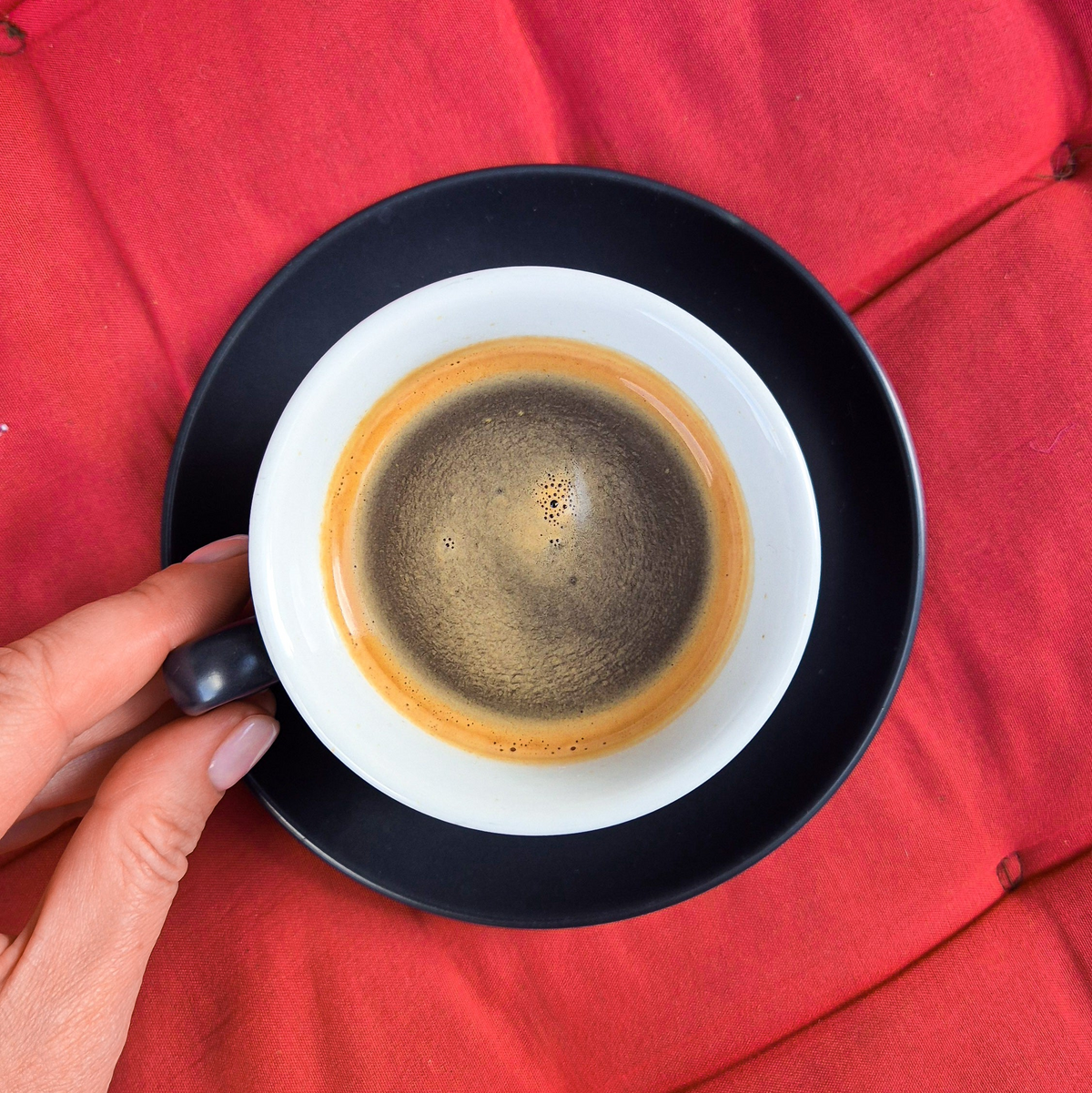A hand holding the handle of a black and white espresso cup with espresso inside, set against a red tufted background.