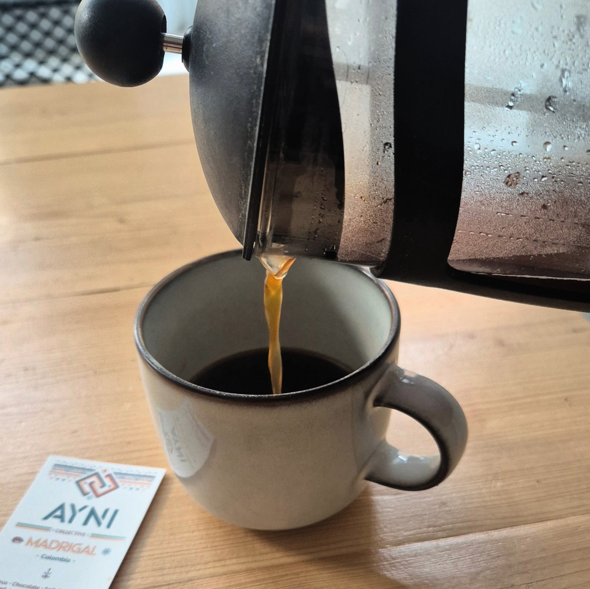 pouring coffee from cafetière into the cup with AYNI product card (Madrigal) on the side