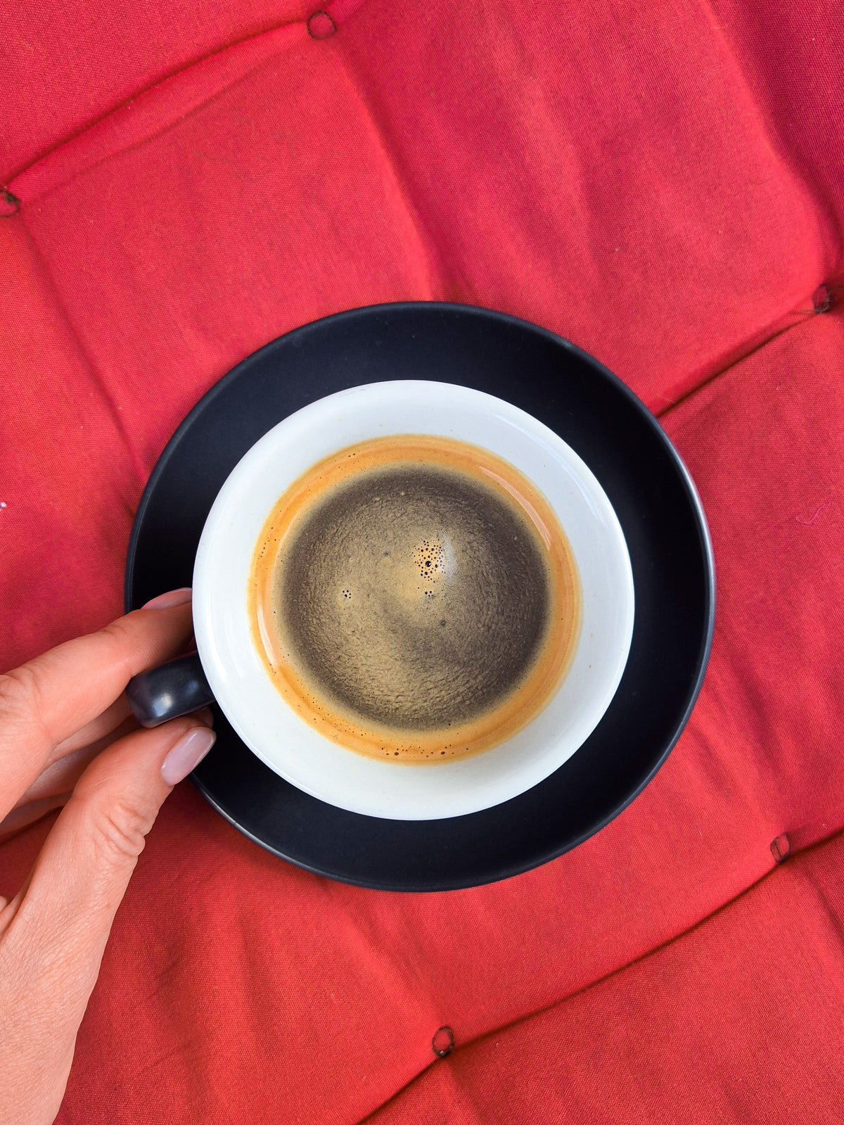 A hand holding the handle of a black and white espresso cup with espresso inside, set against a red tufted background.