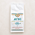 A white bag of Ayni Collective Decaf Luna Colombia medium roast coffee, featuring colorful geometric patterns and teal branding text, set against a light beige textured background.