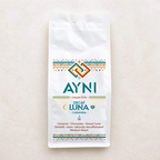 A white bag of Ayni Collective Decaf Luna Colombia medium roast coffee, featuring colorful geometric patterns and teal branding text, set against a light beige textured background.