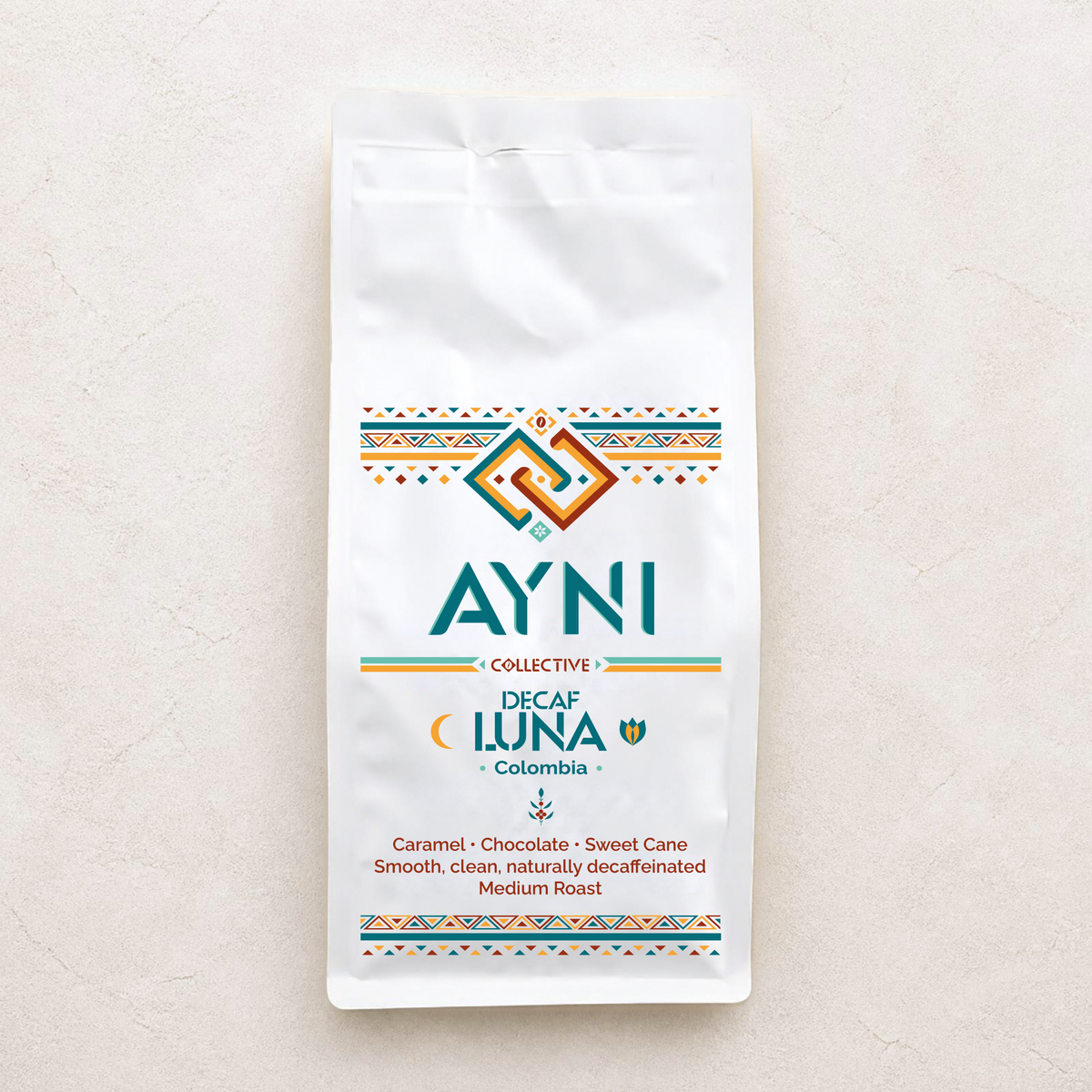 A white bag of Ayni Collective Decaf Luna Colombia medium roast coffee, featuring colorful geometric patterns and teal branding text, set against a light beige textured background.