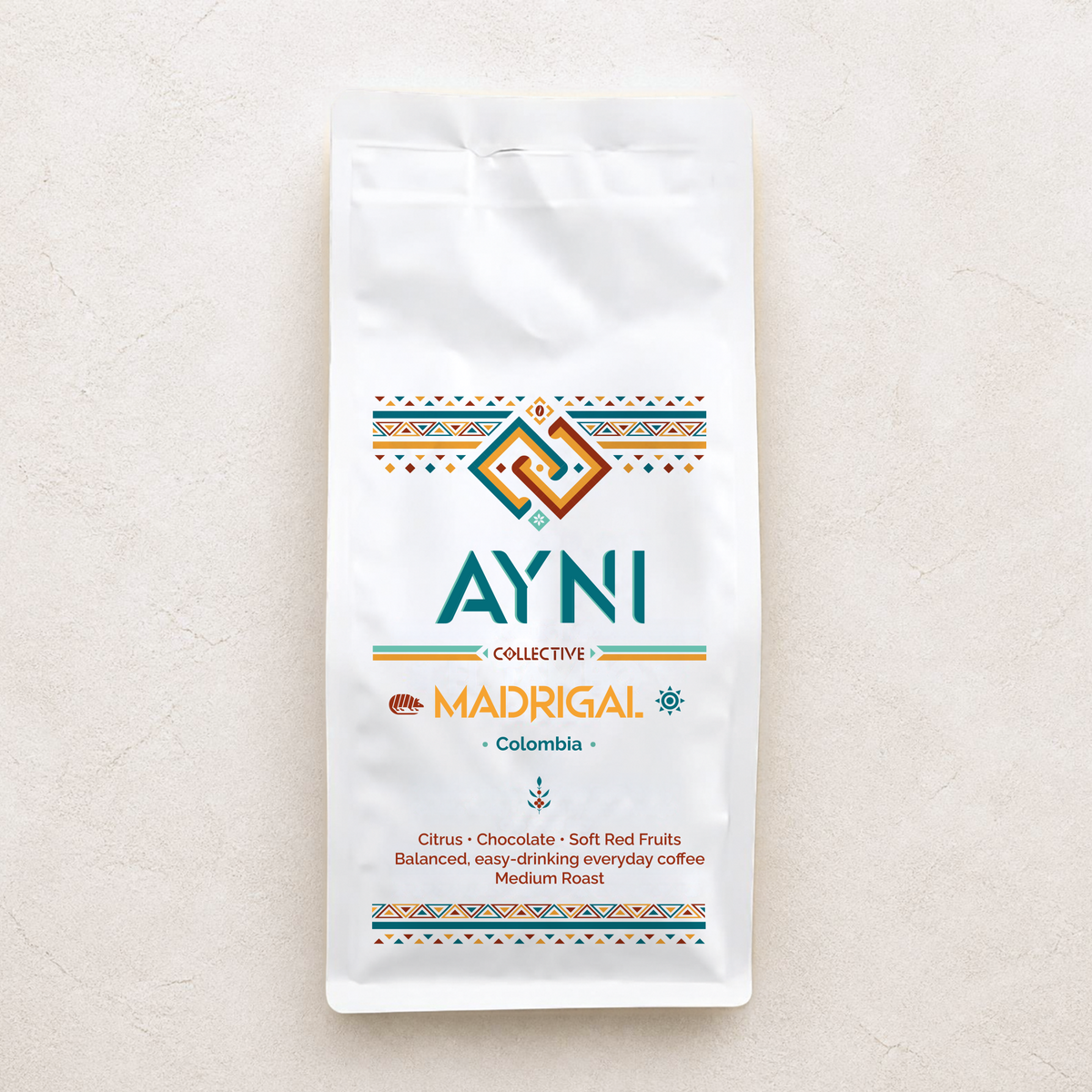 A white bag of Ayni Collective Madrigal Colombia coffee, featuring a colorful geometric logo and product details, set against a textured off-white background.