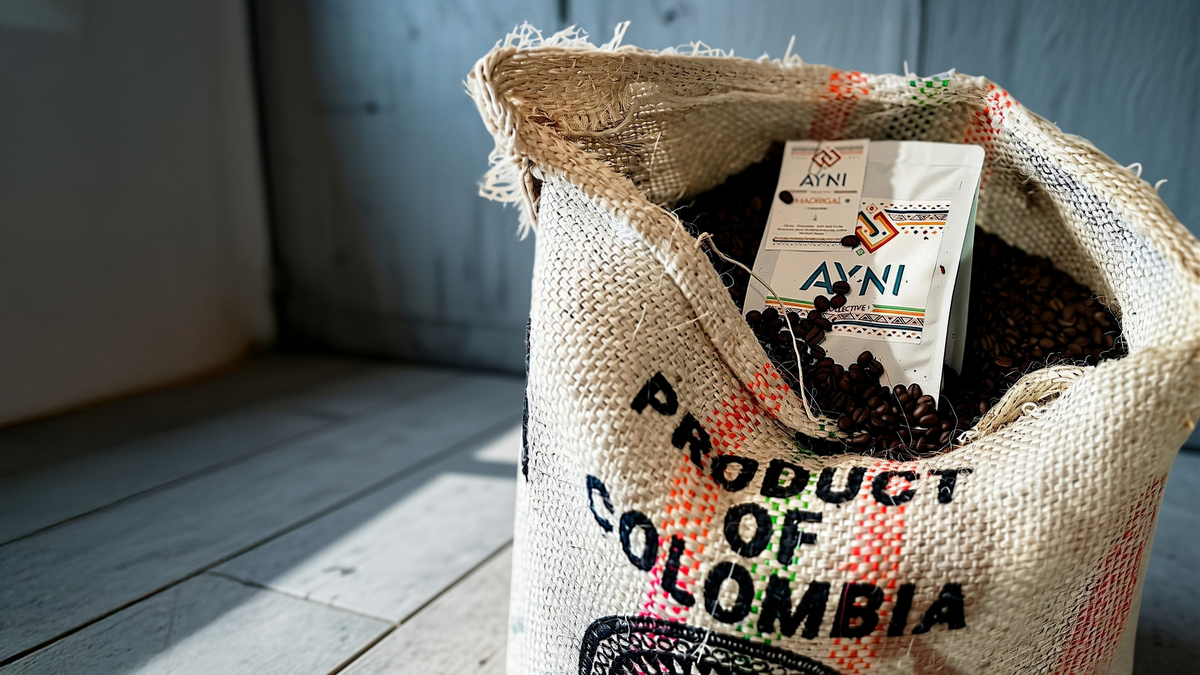 A burlap sack labeled 'PRODUCT OF COLOMBIA' filled with coffee beans and a visible Ayni coffee package, photographed in a rustic indoor setting with wood flooring and a blue-gray wall.